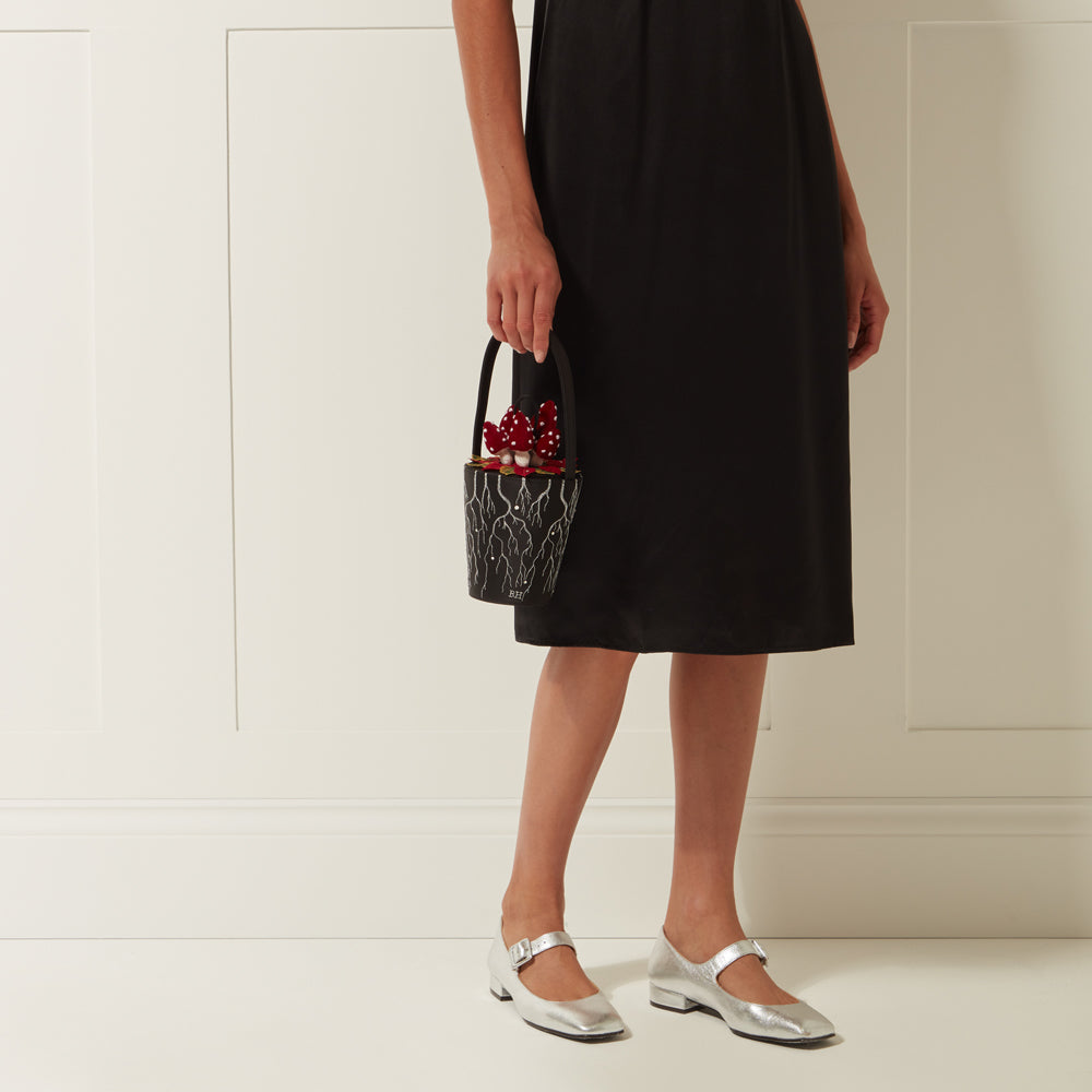 Woman in black dress and silver shoes holding a small black bucket bag with red mushrooms and white lightning bolt design.