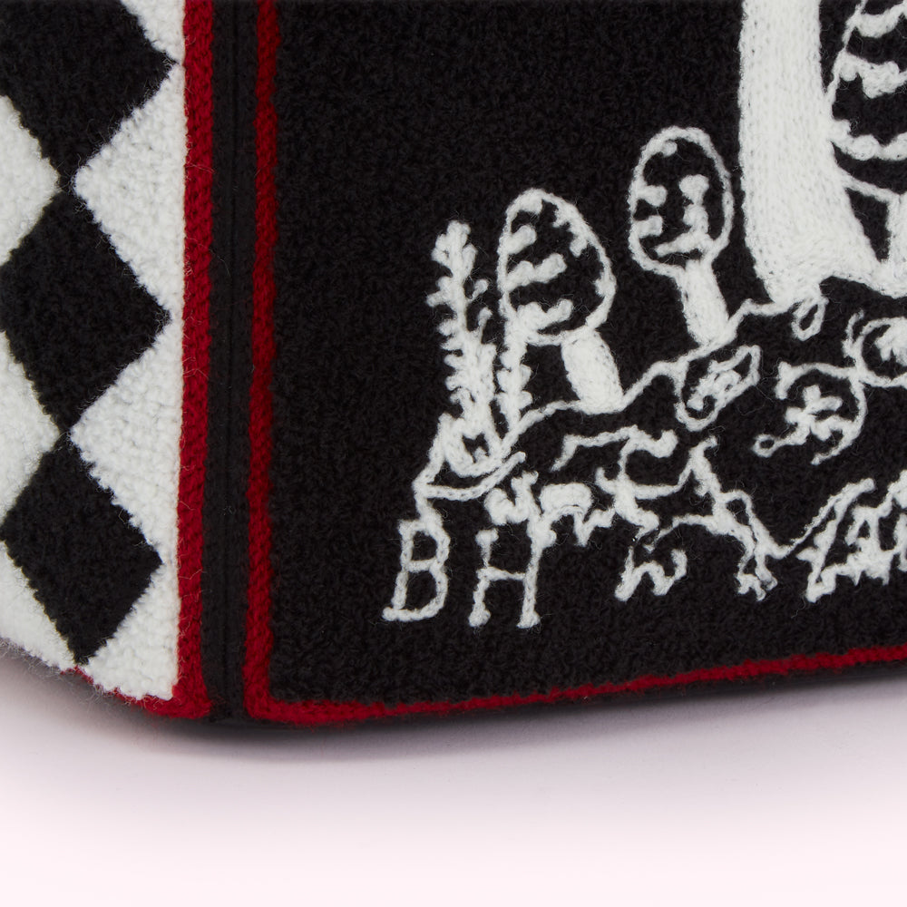 Close-up of a black and white knitted fabric with a red stripe, featuring an embroidered design of mushrooms and foliage, and the initials "BH".
