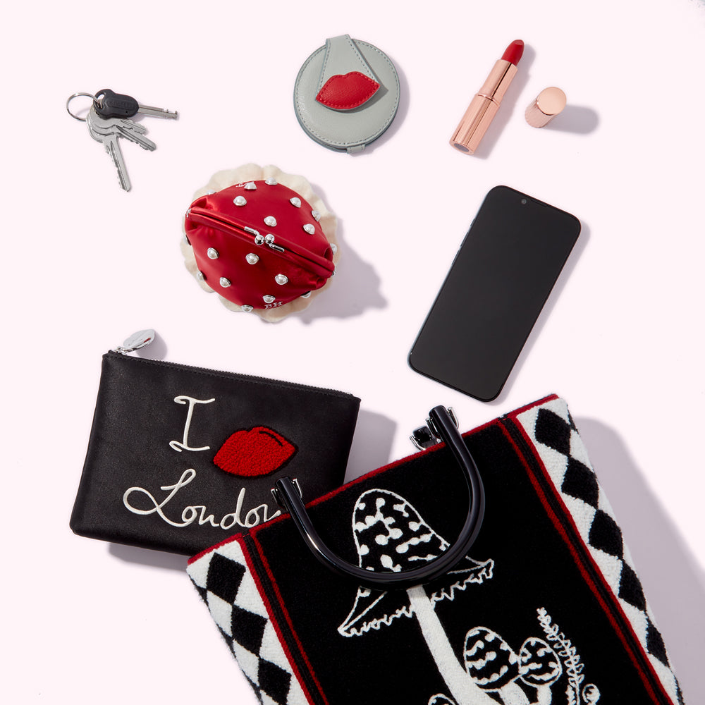 Flatlay of women's accessories on a white background including a handbag with mushroom design, a black pouch with "I love London" embroidered on it, a red coin purse, a compact mirror, lipstick, and keys.
