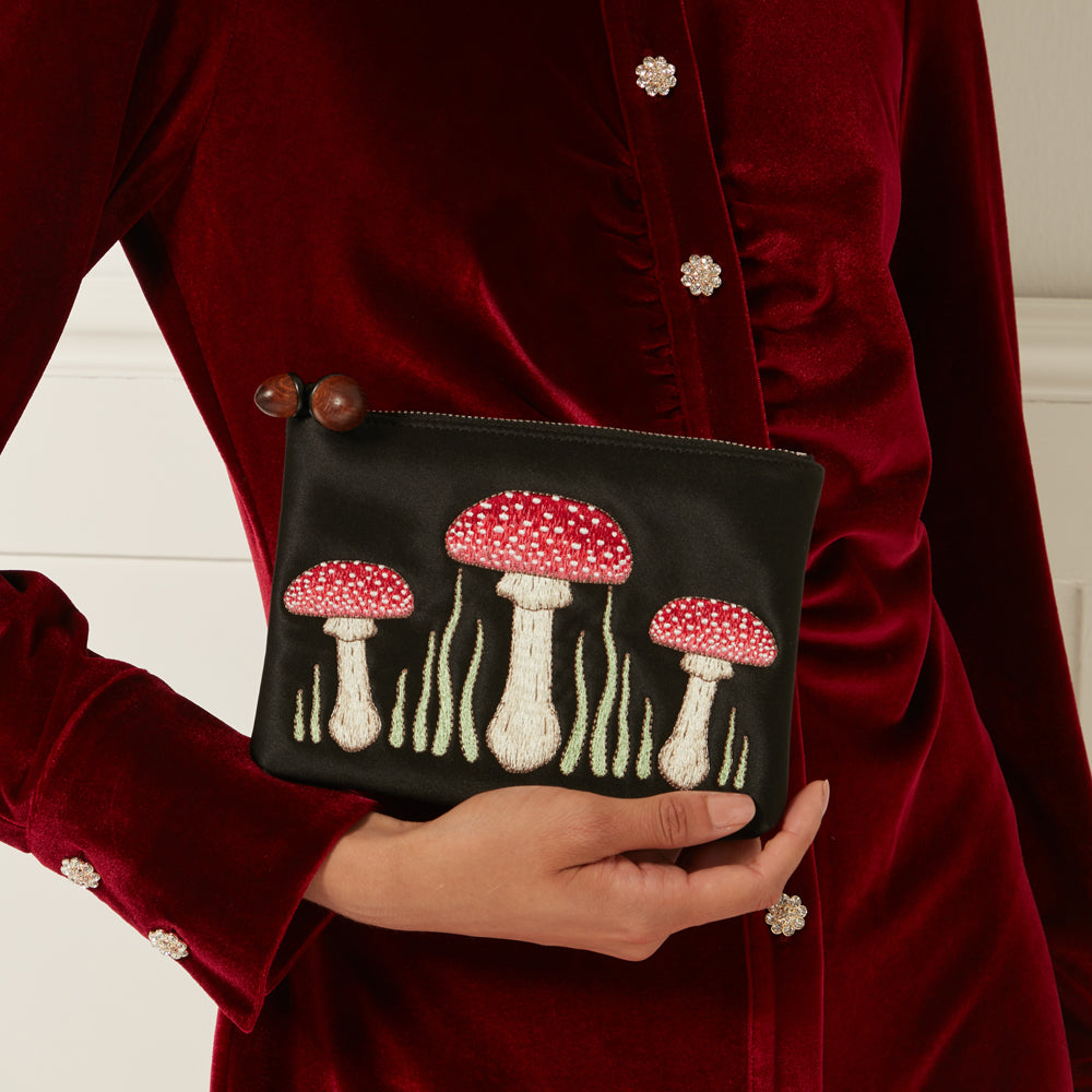 A woman holds a black satin cosmetic bag embroidered with three red and white mushrooms and green grass against a dark red velvet shirt.
