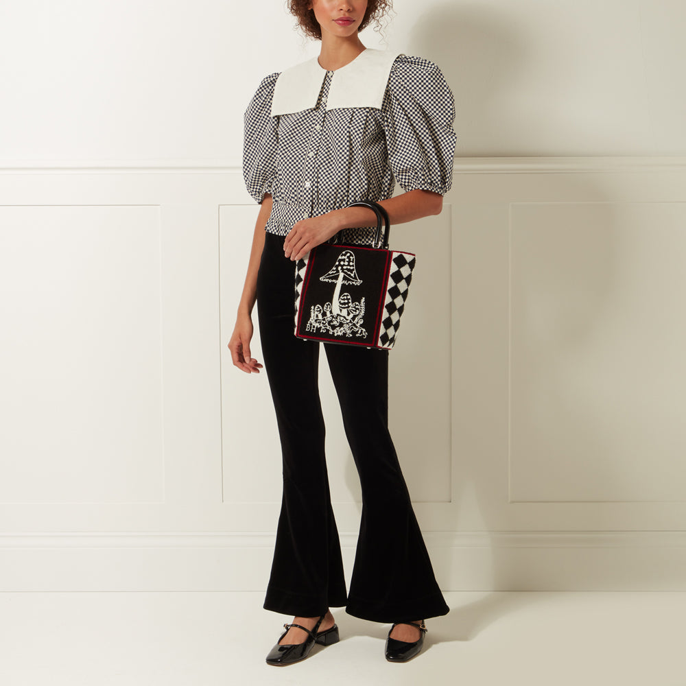 Woman in black pants and white gingham shirt holding a black and white checkered tote bag with a mushroom illustration. The background is white.
