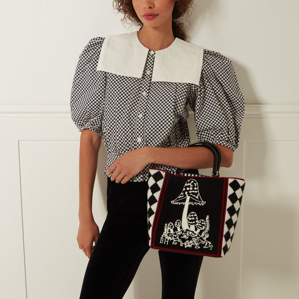 A woman with curly brown hair is modelling a black and white gingham top with a Peter Pan collar and black velvet pants. She is holding a black and white tote bag with a mushroom design. The bag has a black harlequin pattern border and a red band. The background is white.
