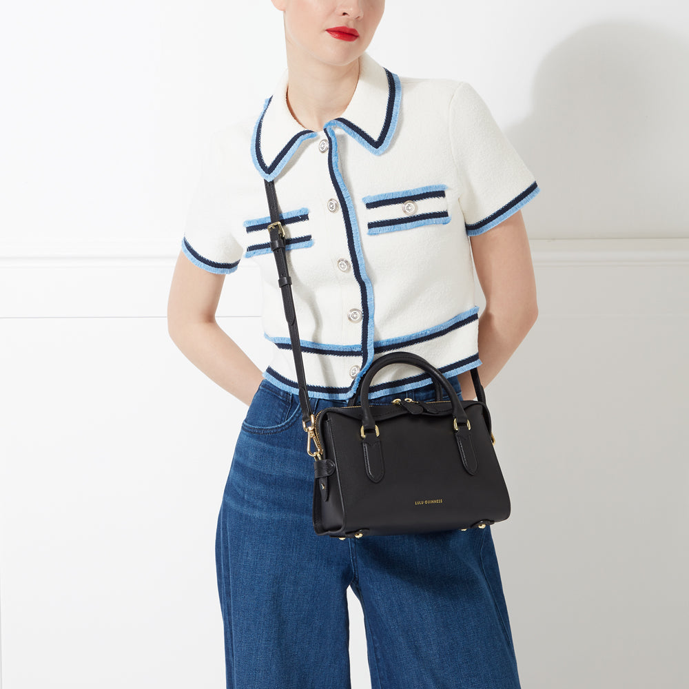 Woman in white sweater and blue jeans carries a black Lulu Guinness crossbody bag.
