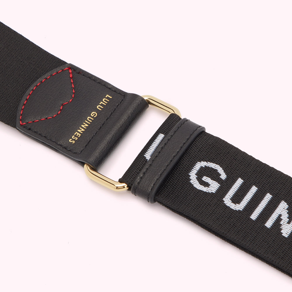 Close-up of a black Lulu Guinness shoulder strap with gold hardware and the brand name embroidered on it. A small black leather heart detail is stitched onto the strap.
