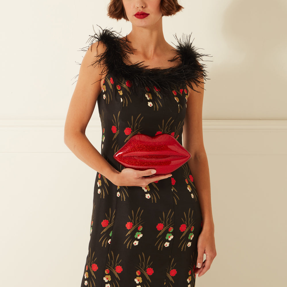 Woman in black floral dress with feather trim neckline holding a red lip-shaped clutch.
