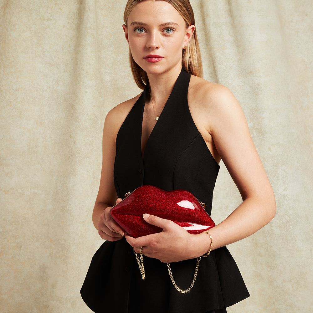 Woman holding a glitter red lip-shaped clutch bag against a beige background