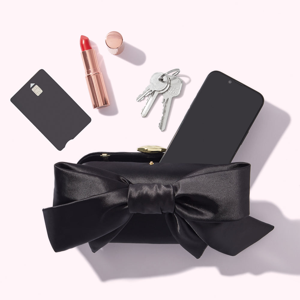 A black satin bow clutch purse containing a smartphone, keys, lipstick, and a credit card on a white background.
