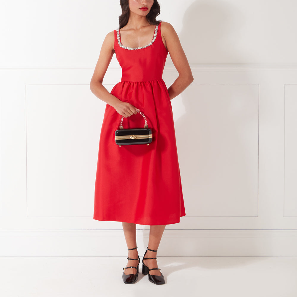 Woman in a red dress and black heels holding a black and gold box purse against a white background.
