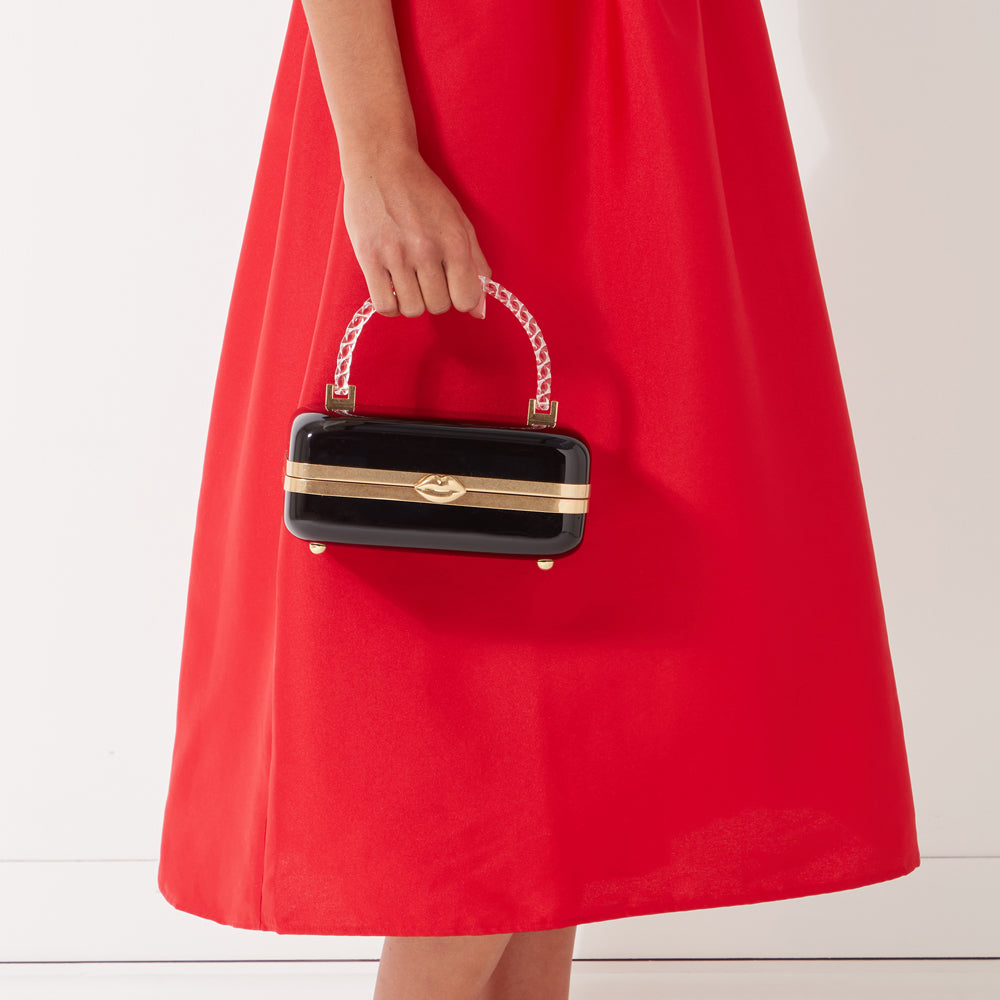 A woman in a red dress holds a black rectangular box clutch with a gold lip detail and a clear handle.
