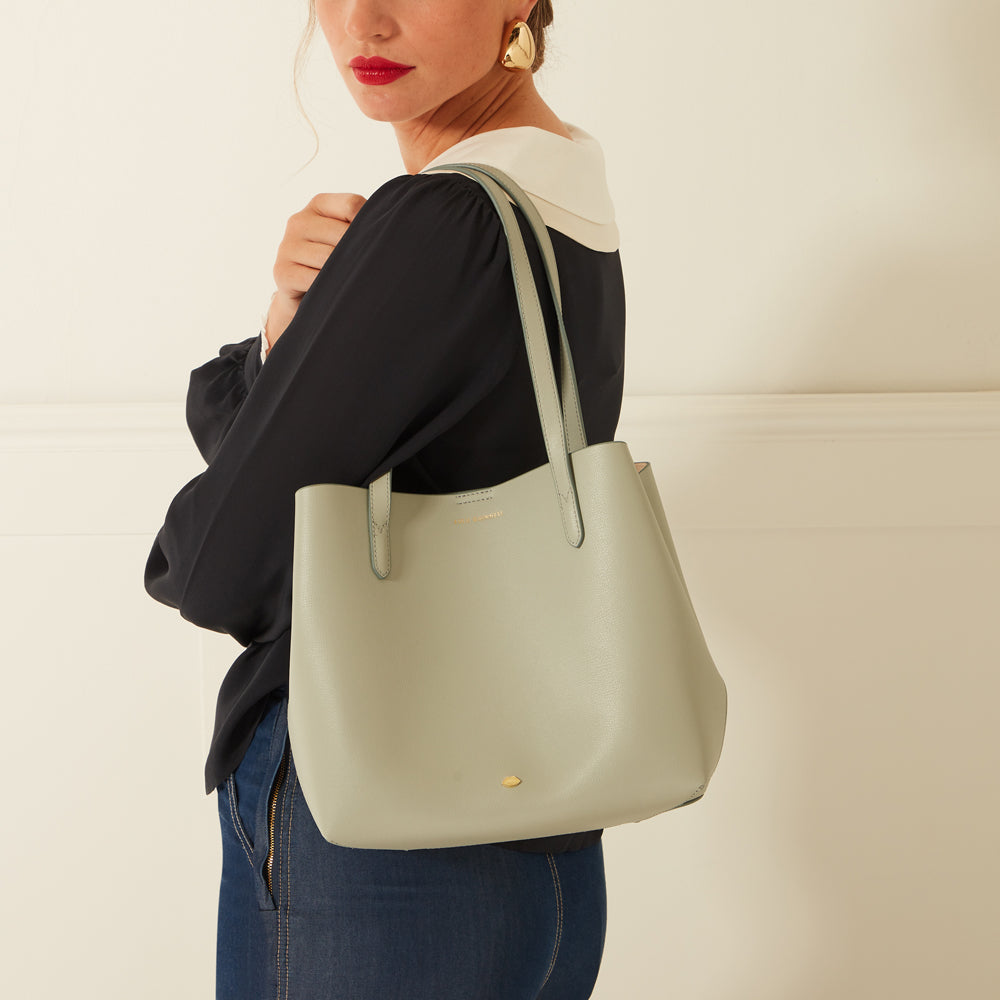 A sage green tote bag held by a woman wearing a black blouse and jeans.
