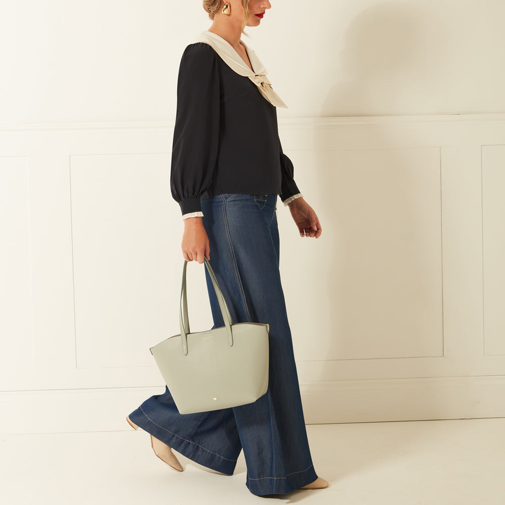 Woman in a black top with a cream collar, dark wash wide leg jeans and sage green tote bag against a white background.
