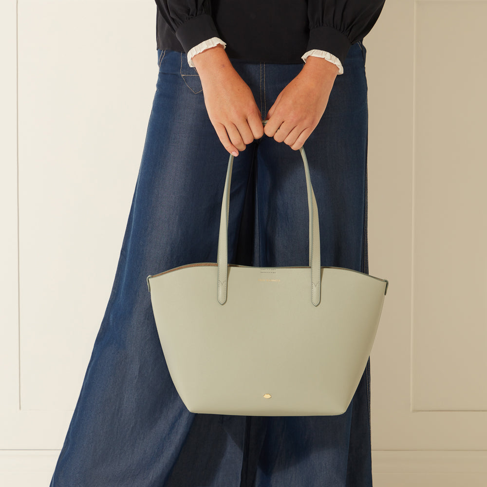 A sage green leather tote bag held by a person wearing dark wash jeans and a black top.

