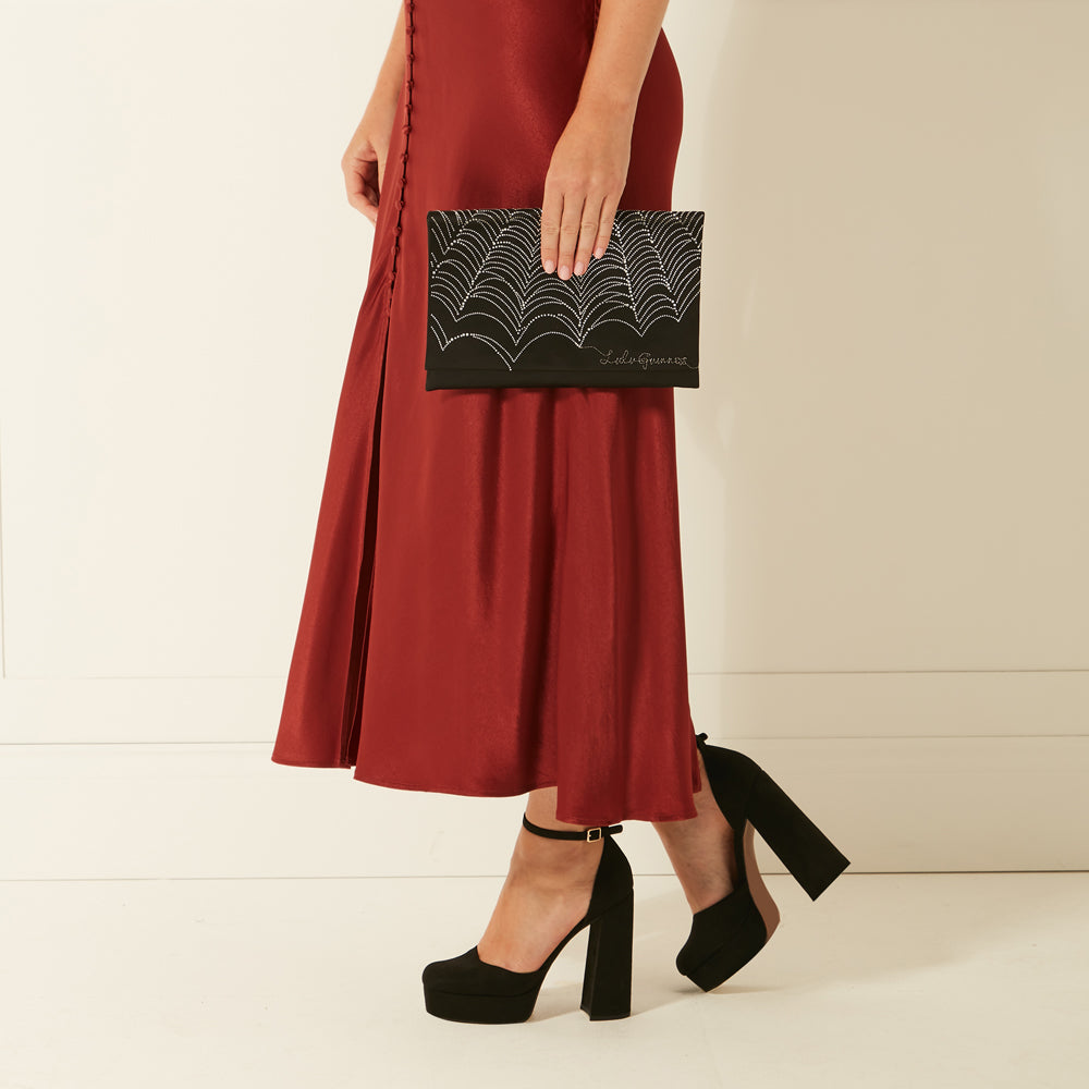 A woman in a rust-coloured midi skirt holds a black clutch with a silver design. She is wearing black platform shoes.