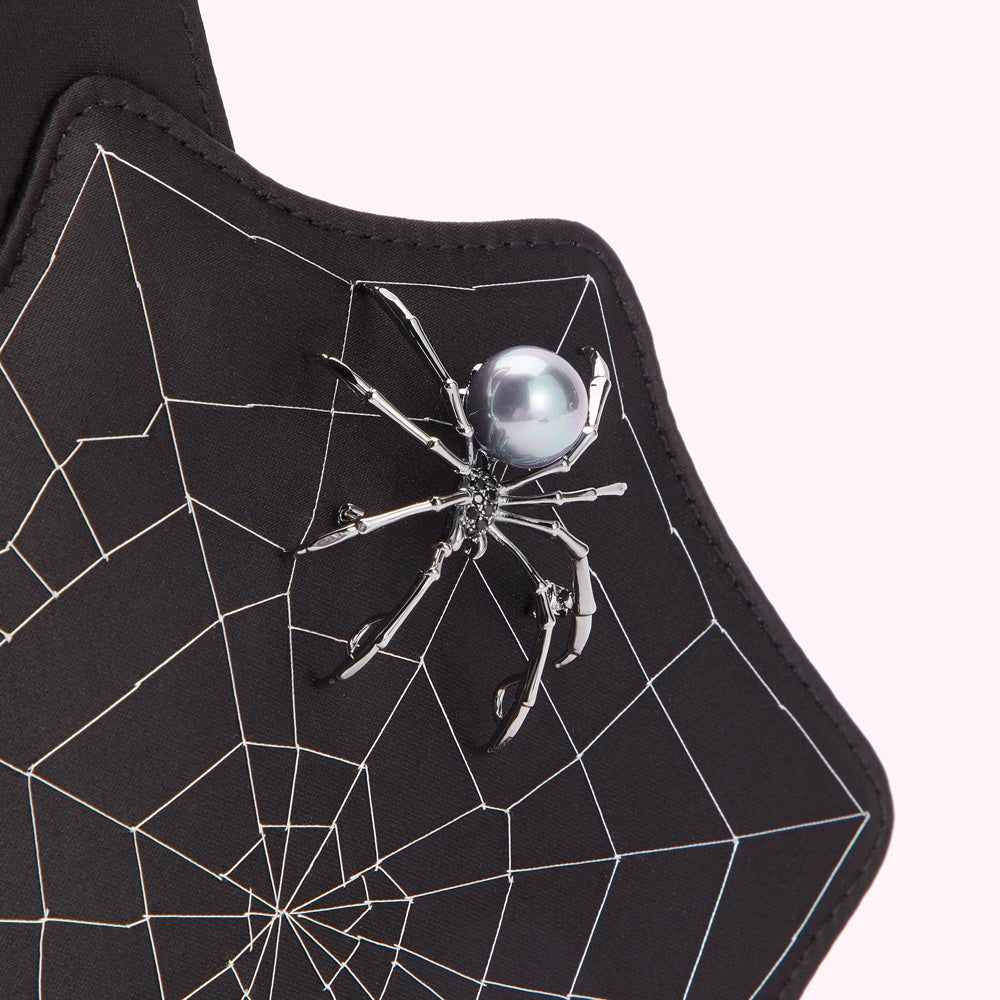 Close-up of a black bag with a spider-shaped brooch featuring a pearl body. The bag is patterned with a spiderweb design.
