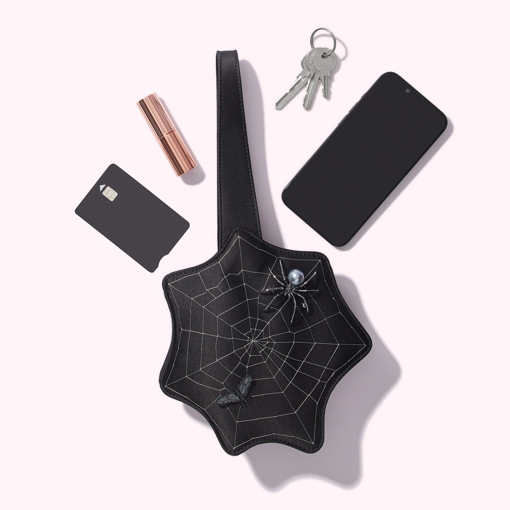 Black spiderweb-shaped purse with lipstick, keys, phone, and credit card on white background.