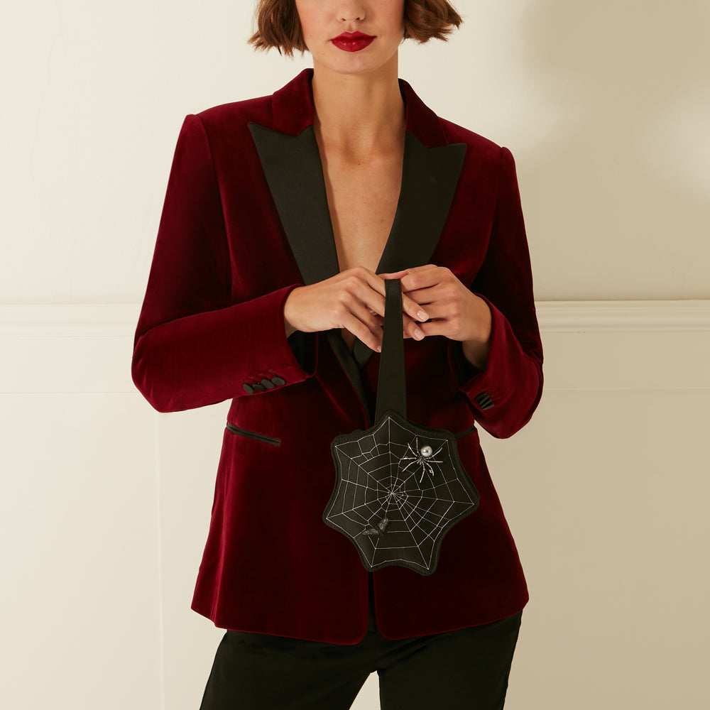Woman in a burgundy velvet blazer holding a black spiderweb-shaped bag.