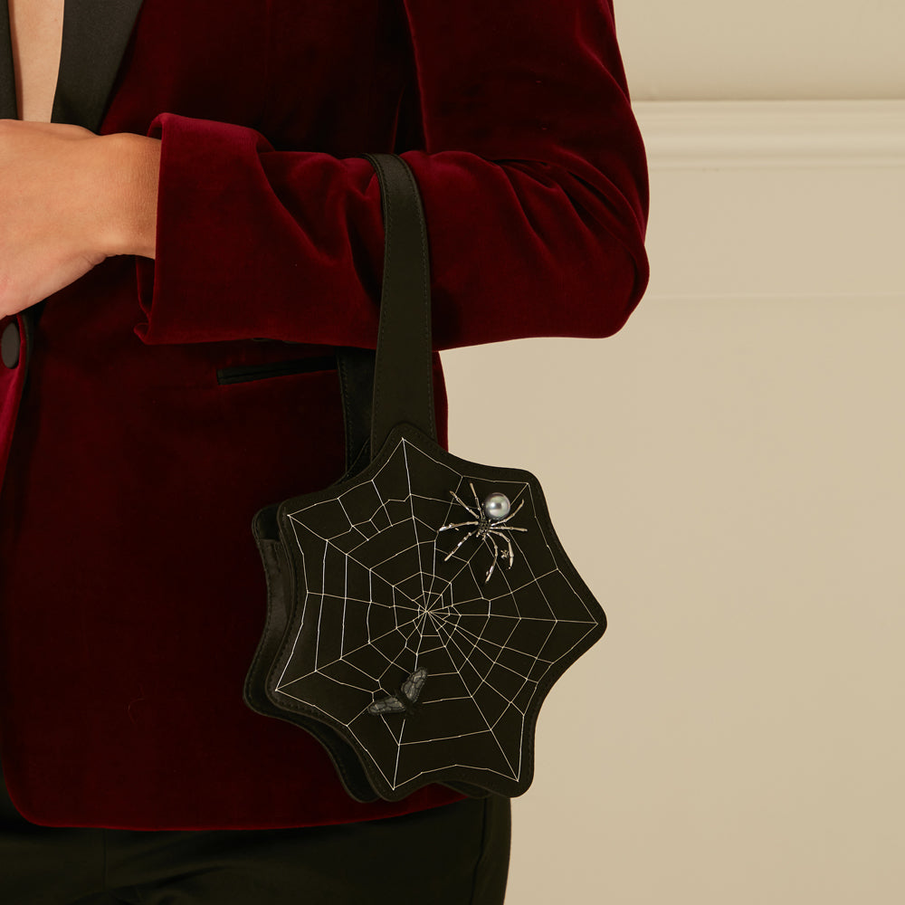 A black star-shaped handbag adorned with a spiderweb design and embellished with a jewelled spider and butterflies, held by a person wearing a burgundy velvet blazer.
