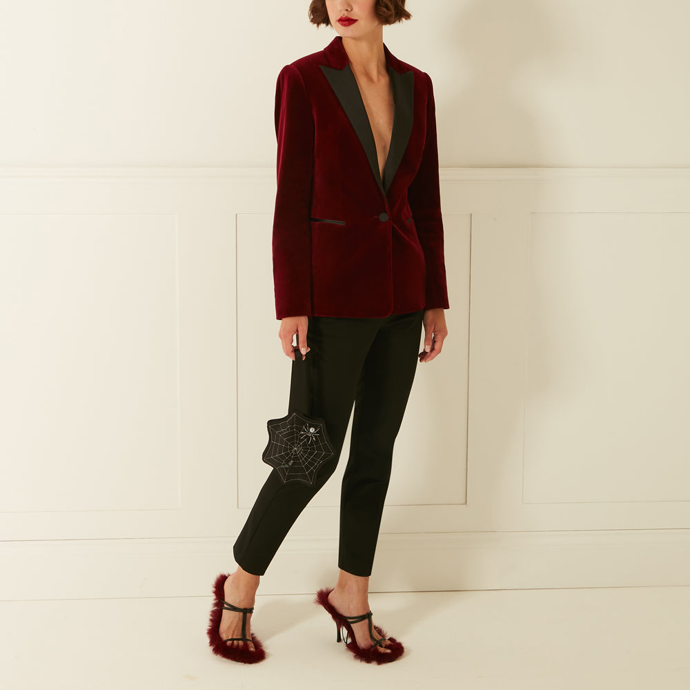 Woman in a burgundy velvet blazer, black pants, and burgundy fuzzy heels holding a black spiderweb-shaped clutch against an off-white background.
