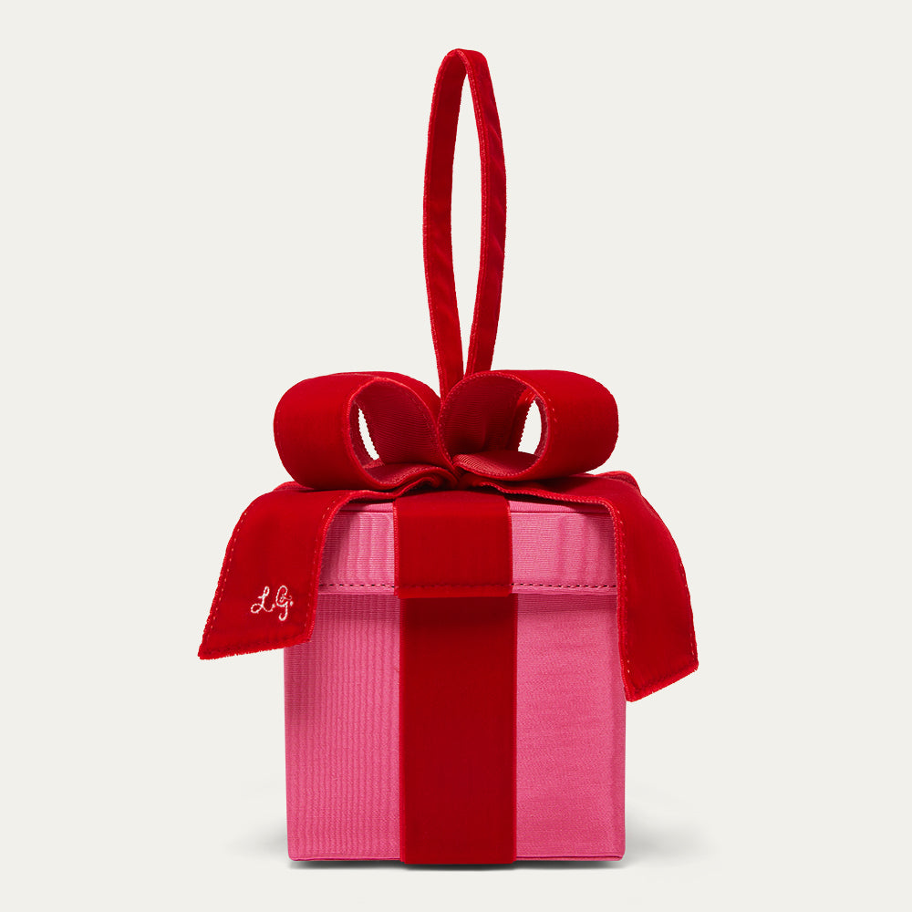 A pink present-shaped handbag with a red velvet ribbon and bow, featuring a top handle and a small embroidered monogram on the ribbon, set against a light gray background.