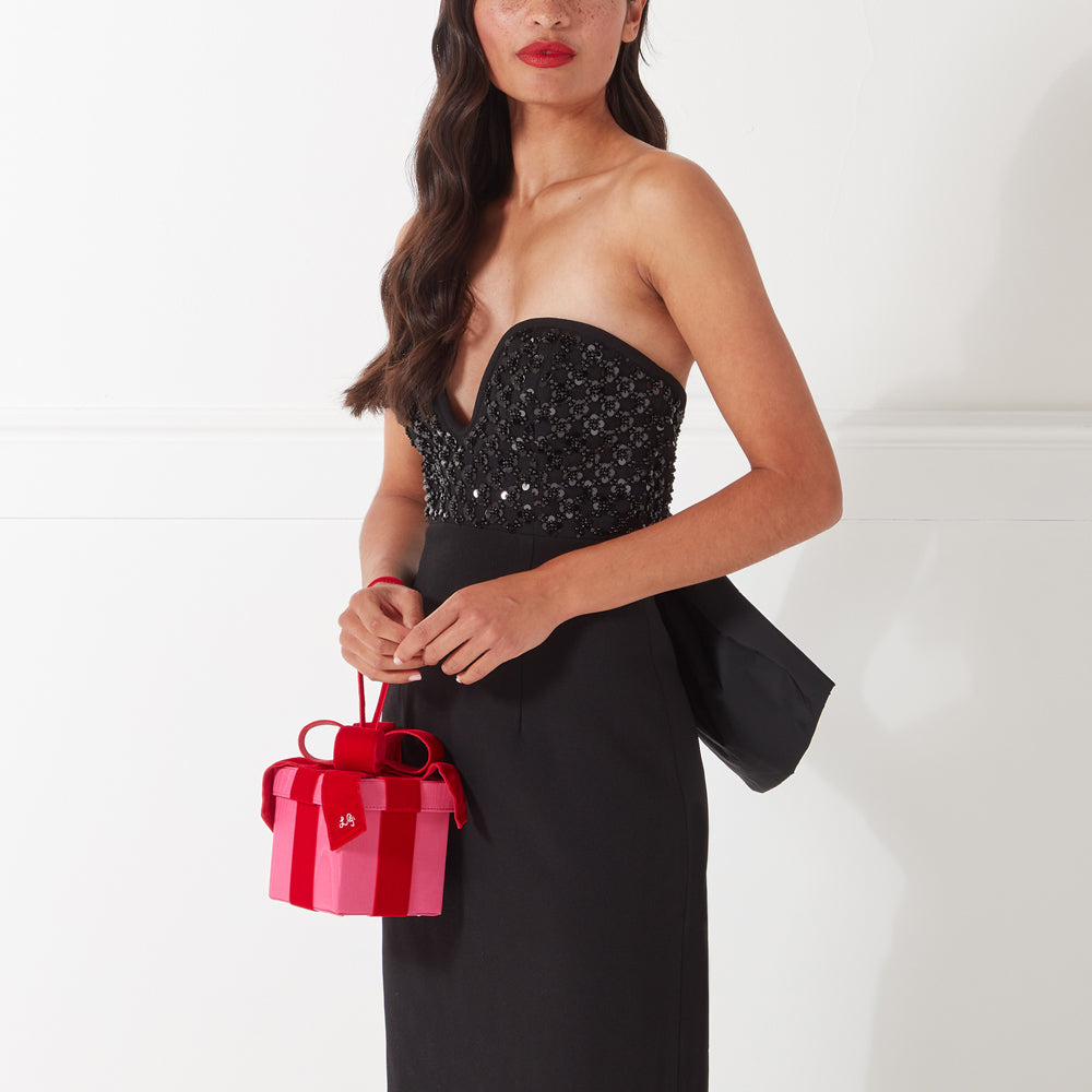 Woman in black beaded strapless dress holding a pink and red velvet gift box-shaped purse against a white background.

