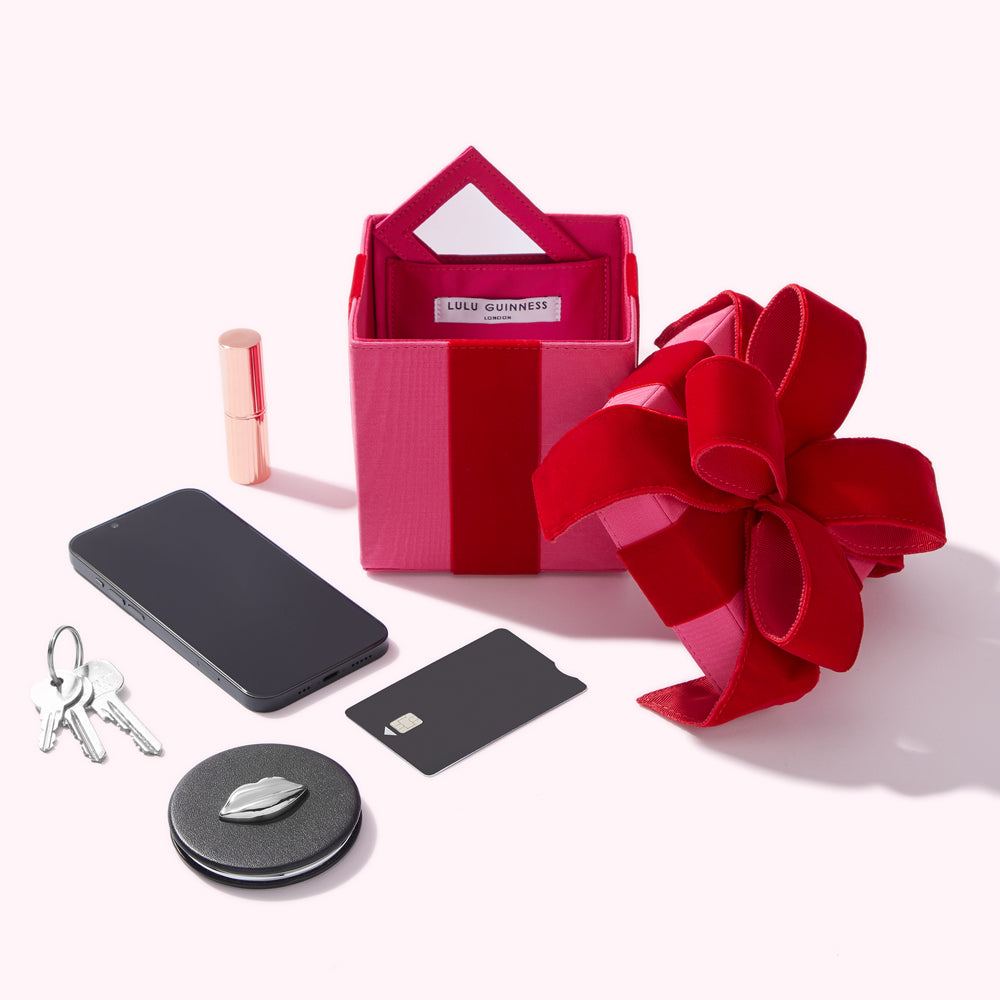 Pink Lulu Guinness gift box with a red ribbon, next to a smartphone, keys, lipstick, and a credit card, all on a white background.
