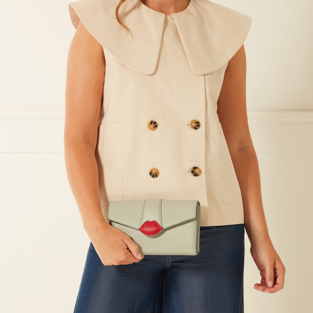 Woman in a beige sleeveless collared vest holding a light green clutch with a red lip design. The background is off-white.
