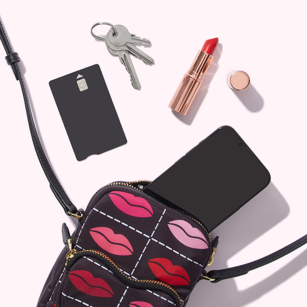 A black crossbody bag with a lip print design containing a smartphone, credit card, keys and lipstick on a white background.