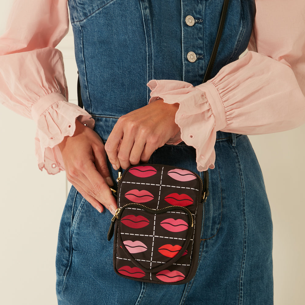 A person wearing a denim dress and a pink blouse is shown holding a black crossbody bag with a repeating pattern of lips and a heart.