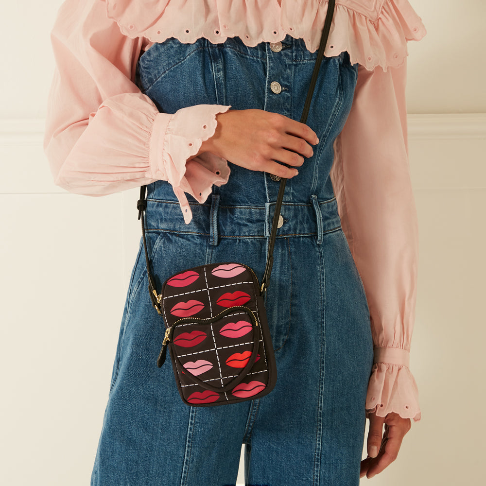 A dark brown crossbody bag with a pink and red lips pattern worn over a denim jumpsuit and a pink eyelet blouse.