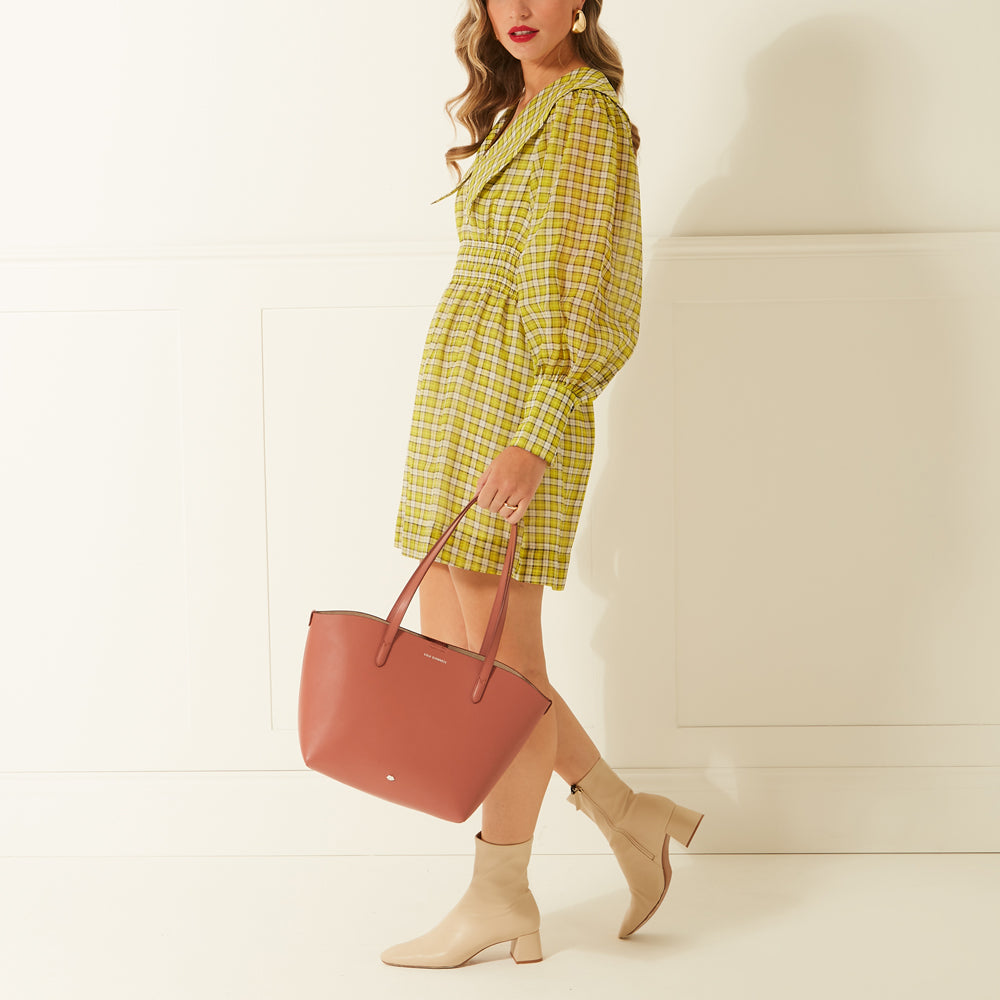 Woman in a yellow gingham dress and beige boots carrying a dusty rose tote bag against a white background.
