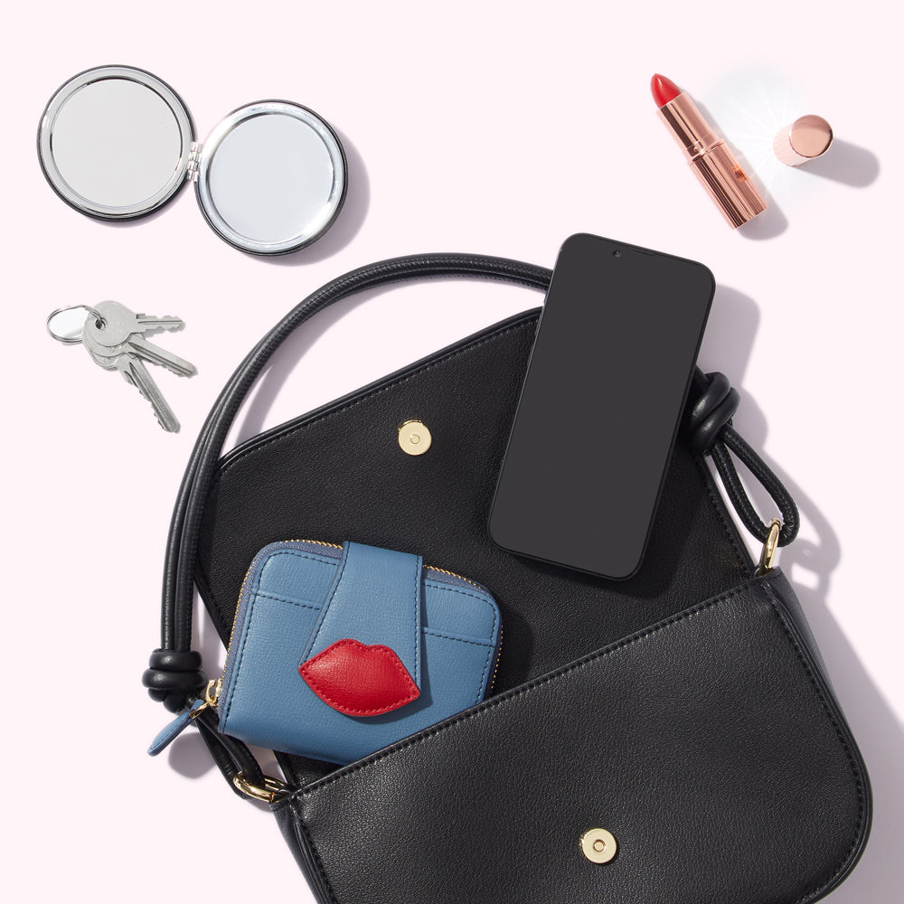 A black handbag with a blue wallet, a smartphone, keys, a compact mirror, lipstick, and lip gloss inside. The background is light pink.
