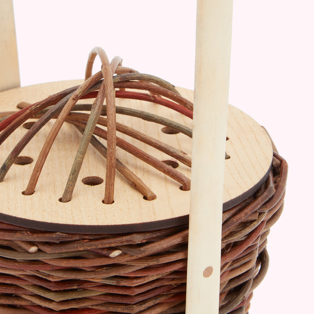 Close-up of a small, round wicker basket with a wooden top featuring thin, multi-coloured twigs woven into a pattern.