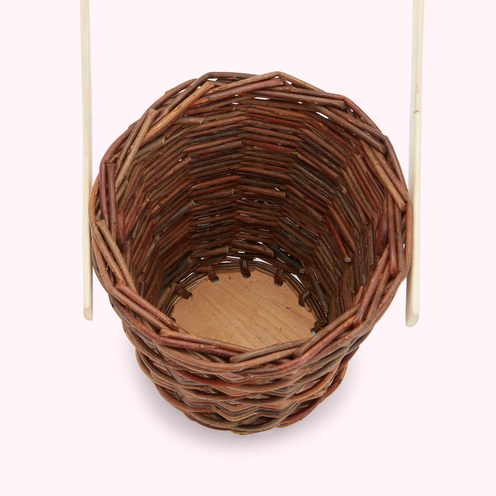 Close-up of a woven brown wicker basket with a round wooden base, displayed against a light pink background, with white handle supports visible on either side.