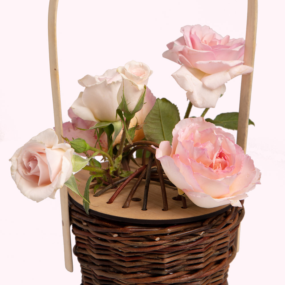A small wicker basket with a light wooden handle holds a bouquet of light pink roses.