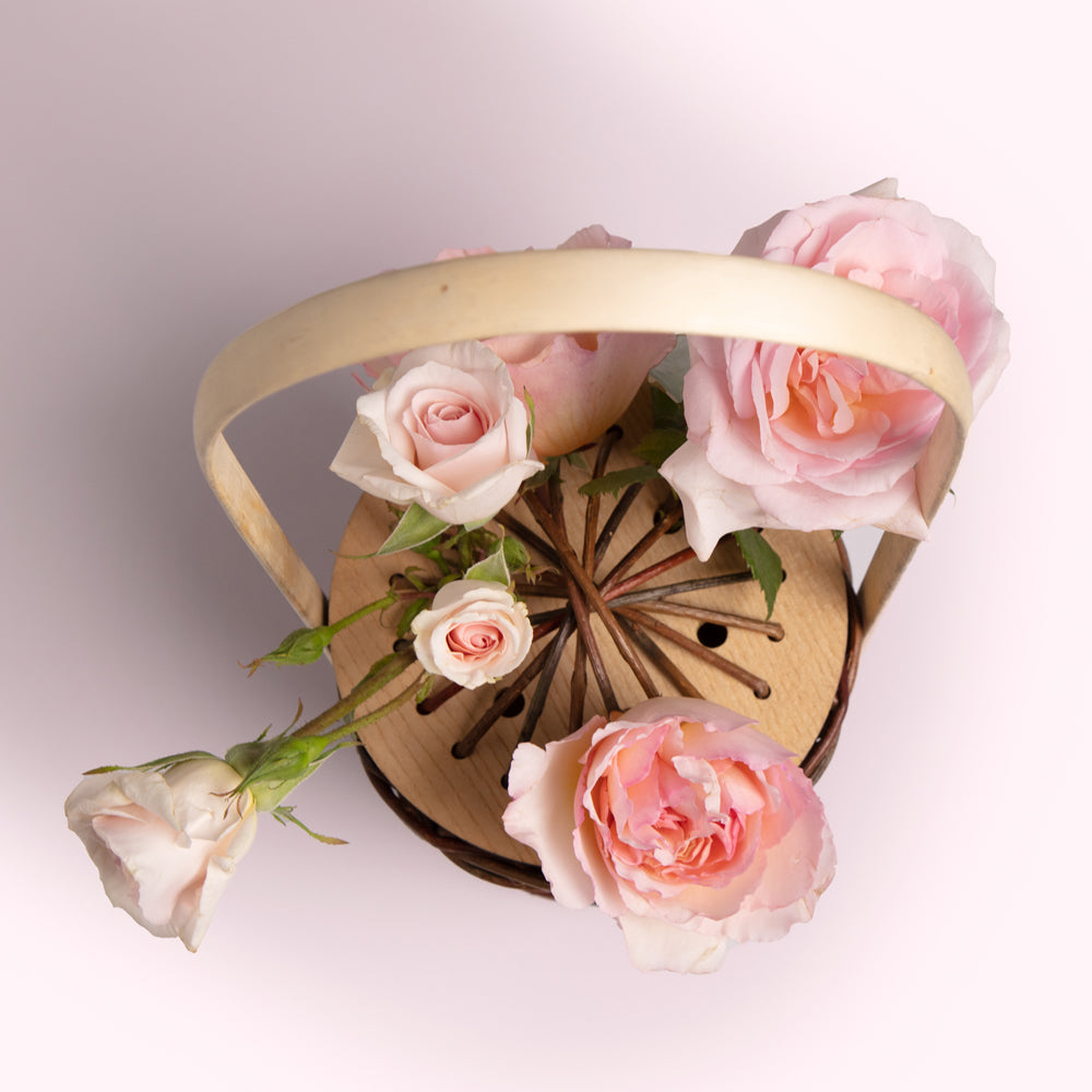 Overhead view of a light wooden basket filled with several light pink roses against a light pink background.

