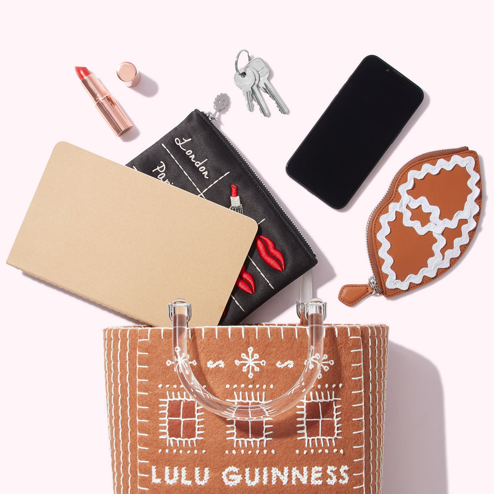 A flat lay shot against a white background of a Lulu Guinness gingerbread house-shaped tote bag with its contents spilling out: a notebook, a black embroidered makeup bag, a smartphone, keys, lipstick, and a lip-shaped coin purse.
