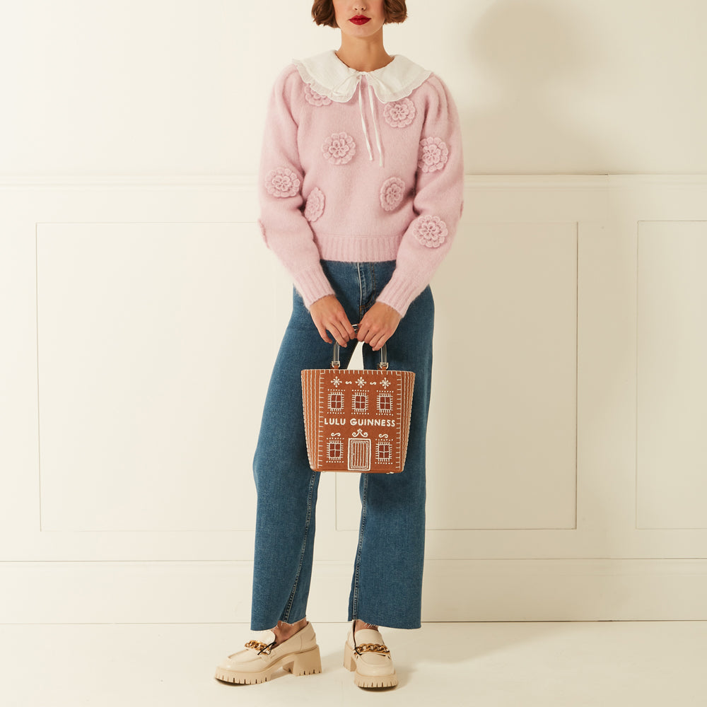 Woman in a pink sweater and jeans holding a brown Lulu Guinness gingerbread house tote bag against a white background.
