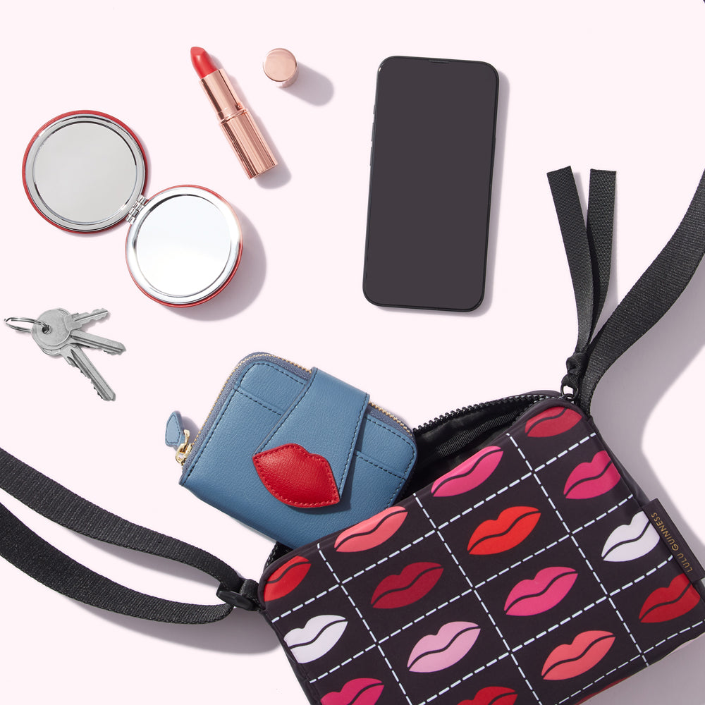 A black crossbody bag with a red lips pattern, a blue wallet, keys, a compact mirror, lipstick and a smartphone on a white background.
