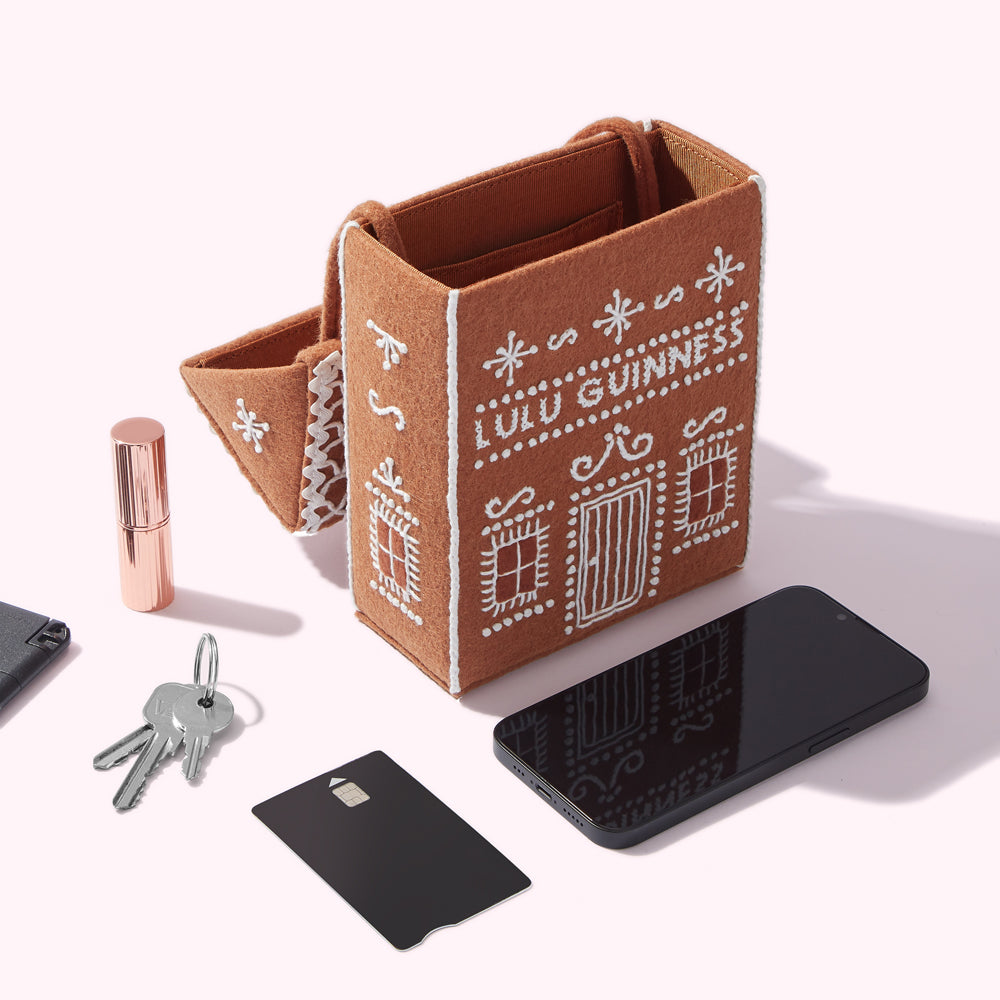A brown felt Lulu Guinness gingerbread house-shaped bag sits on a white surface next to a smartphone, keys, a credit card, and lipstick.
