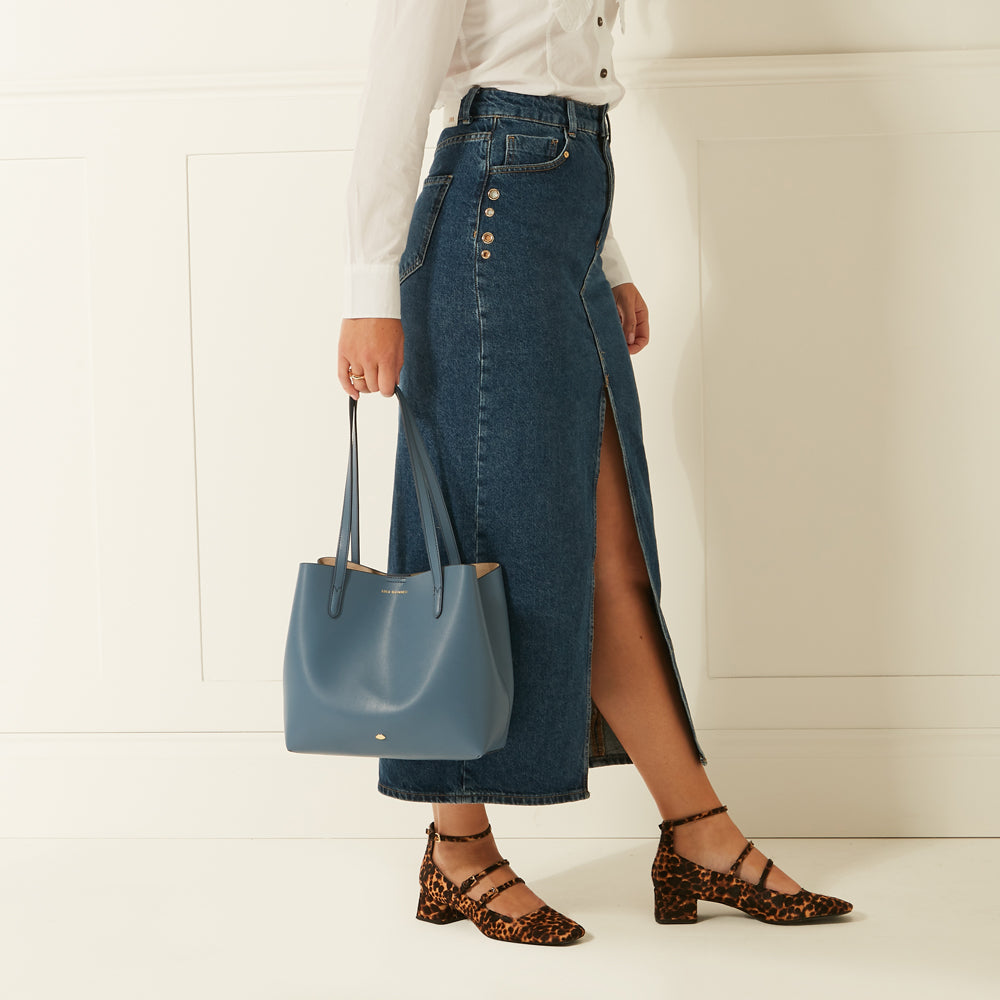 Woman in white shirt and denim maxi skirt carries a blue tote bag and wears leopard print shoes.