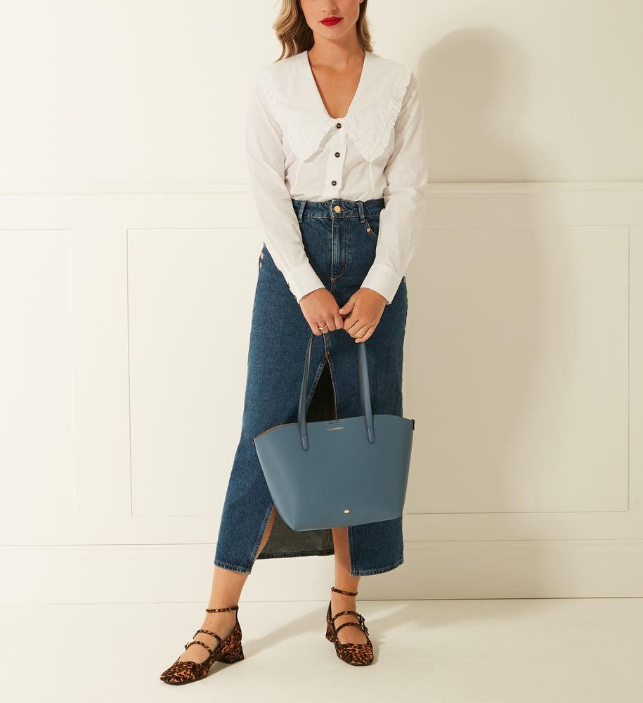 A woman in a white Peter Pan collar blouse and denim midi skirt carries a slate blue tote bag against a white background.
