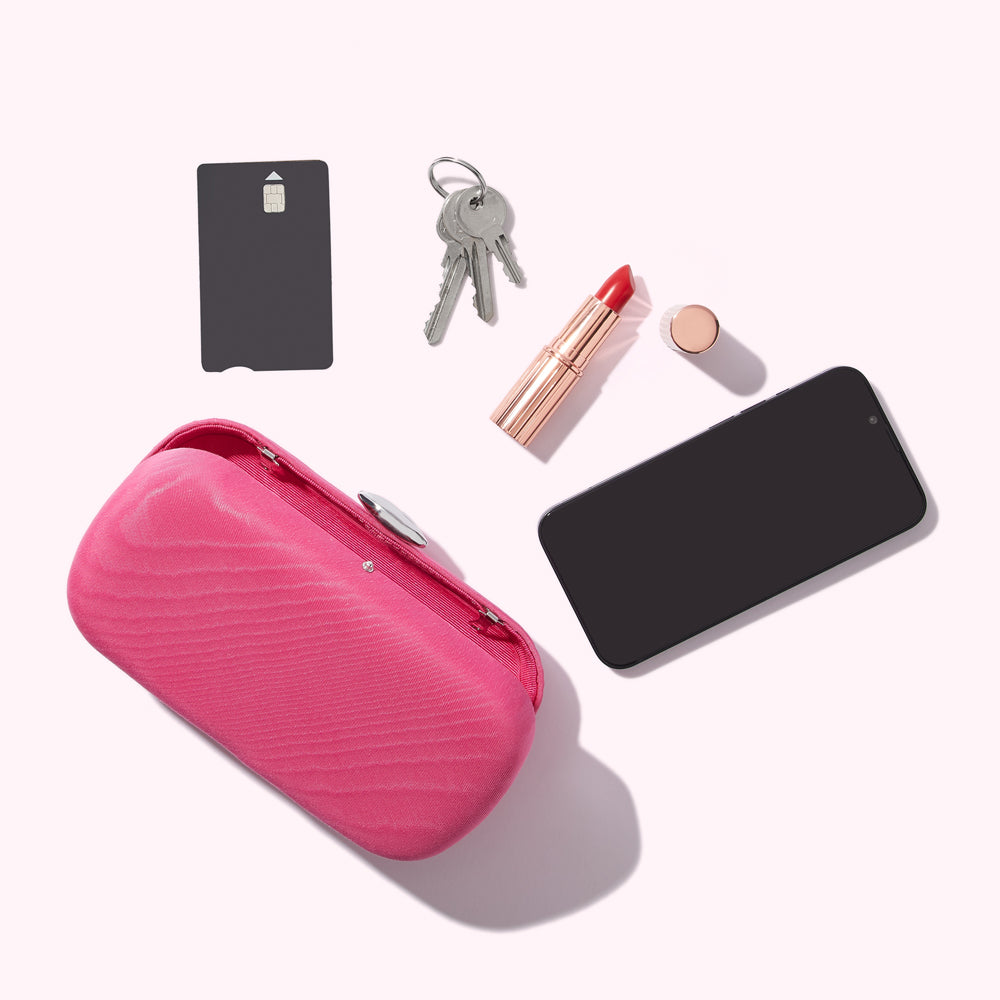 A flat lay image of a pink clutch, a black credit card, keys, lipstick, and a smartphone on a white background.