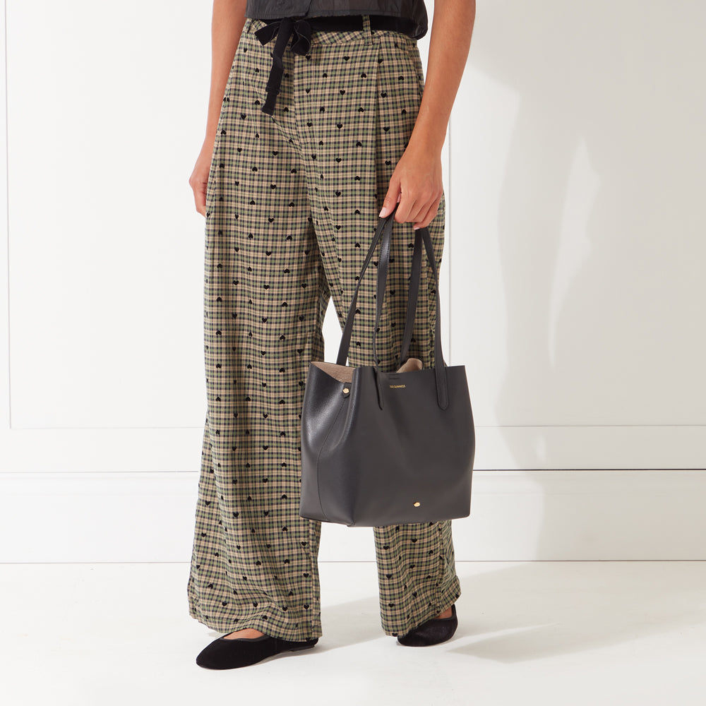 A woman in olive green pants with black hearts patterned on it, holding a black tote bag.