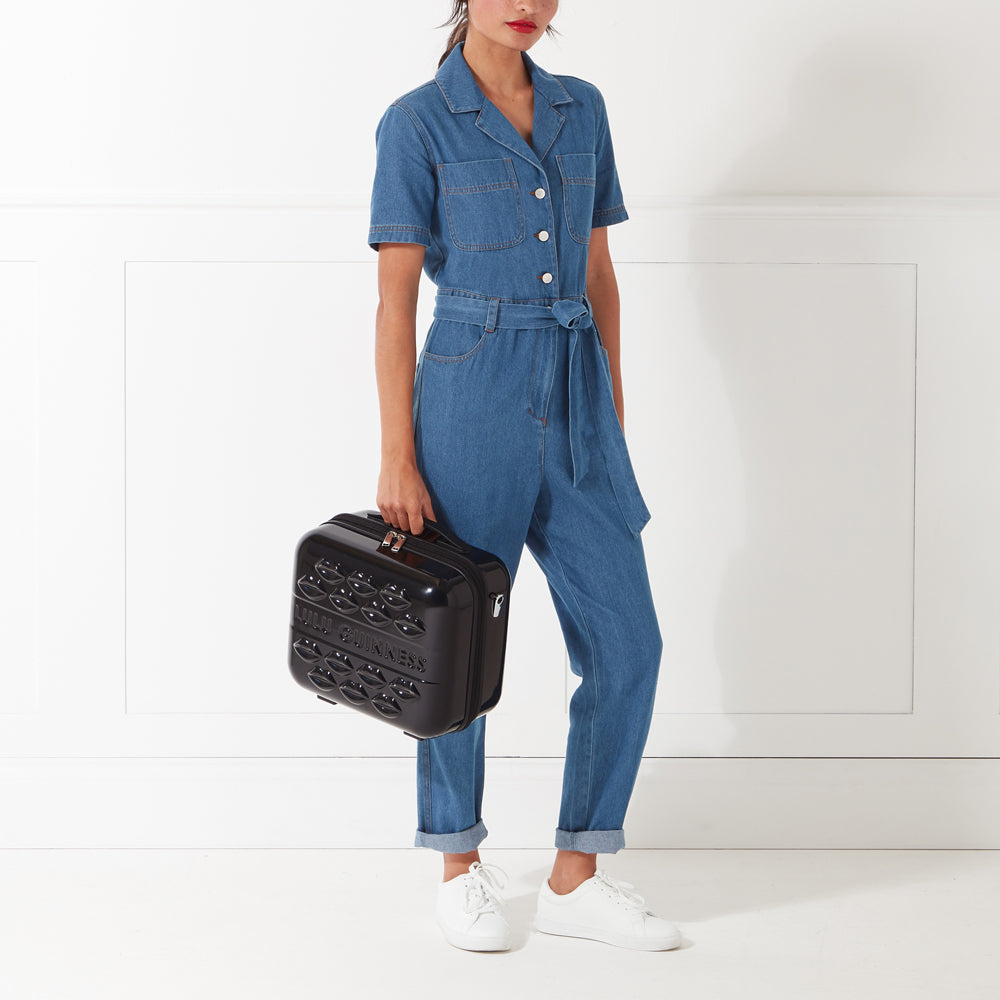 A woman in a denim jumpsuit holds a black Lulu Guinness makeup case against a white background.

