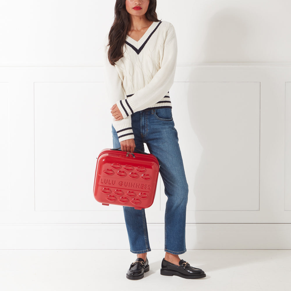 A woman in a cream sweater and jeans holds a red Lulu Guinness makeup case against a white background.

