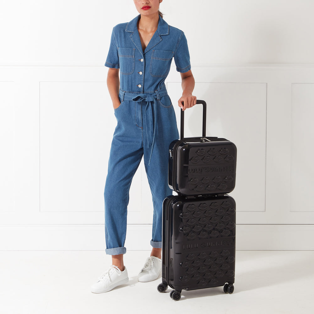 A woman in a denim jumpsuit stands beside a black Lulu Guinness two-piece luggage set featuring a lip print against a white background.
