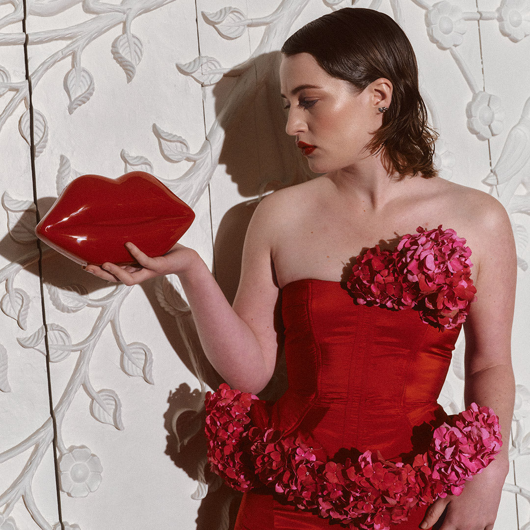 Woman in a red and pink floral dress holding a red lip-shaped clutch bag against a decorative wall.