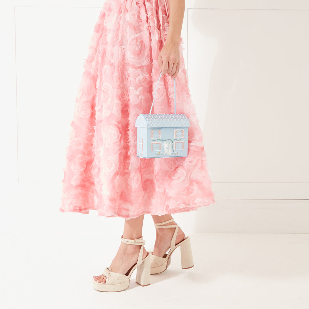 A model wearing a pink dress and cream coloured heeled shoes holding a blue satin house shaped bag down in her hand.