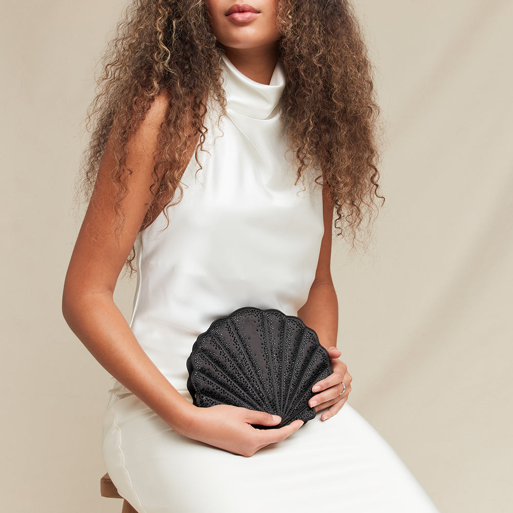 Woman holding a black shell-shaped clutch against a neutral background