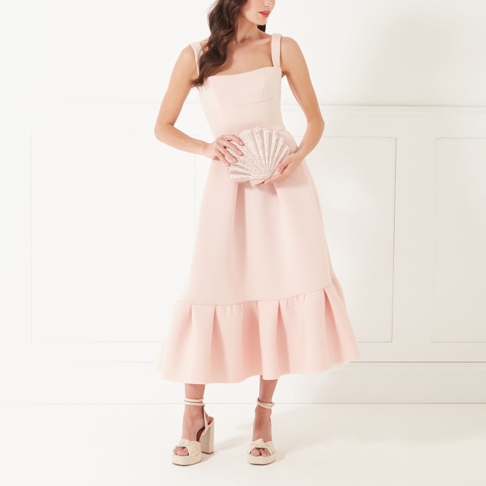 A model with dark hair wearing a pale pink dress and heels holding a blush pink satin shell shaped clutch bag.